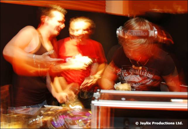 Three blurred people crowd behind DJ decks, one wearing Technics headphones