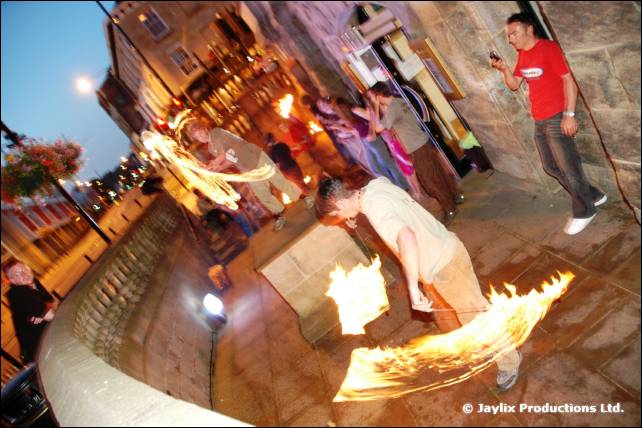 A person swings fire along a cobbled street at dusk, with onlookers and a person in a red shirt