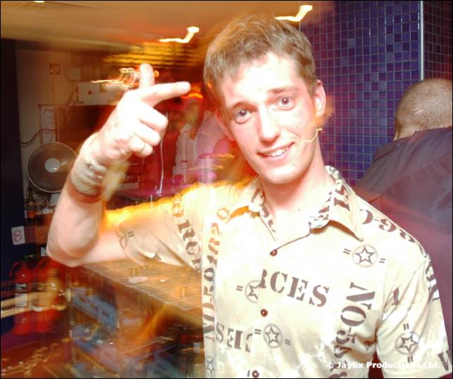 A smiling person in a patterned shirt points at the camera in front of a bar