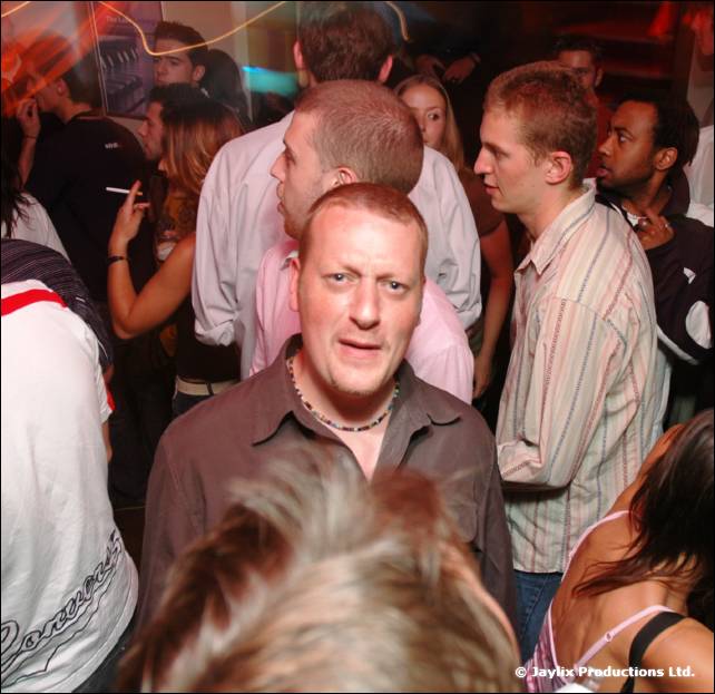 A crowded bar with a man in a brown shirt looking at the camera in the foreground