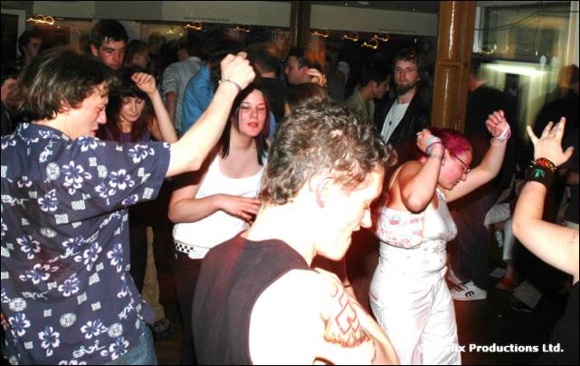 People dance in a busy bar, with a person in a blue patterned shirt in front