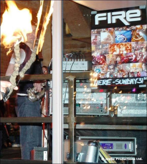 A person with a burning torch beside a window with a FIRE poster and sound equipment