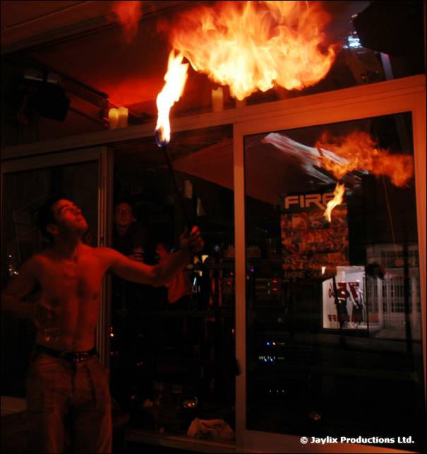 A shirtless fire breather blows a large flame upwards outside a bar window with a FIRE poster