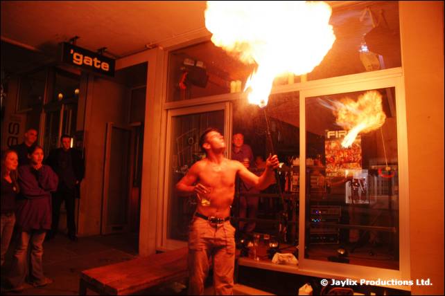 A shirtless person blows a fireball upwards outside a bar window, with a gate sign above