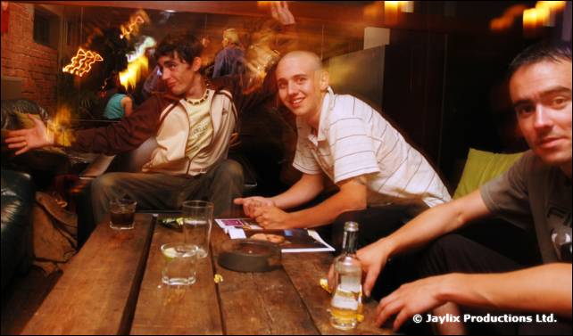 Three men sit at a wooden table with drinks and a bottle in a bar, one leaning in from the right