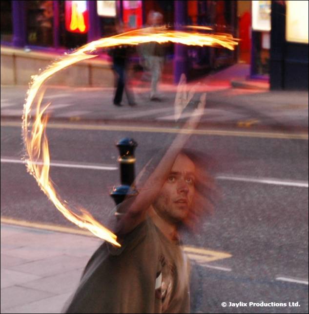 A person on a street swings a fiery arc of poi, looking up