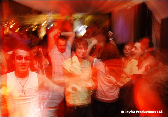 A blurred crowd dances, with a person in sunglasses and a white vest at the front