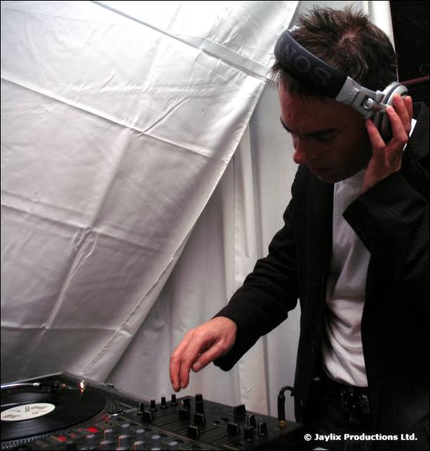 A DJ in headphones and a dark jacket mixes at a turntable inside a white tent