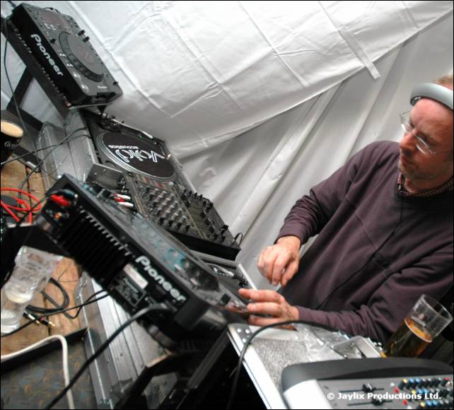 A man in a dark jumper operates Pioneer DJ equipment inside a white tent, a pint beside him