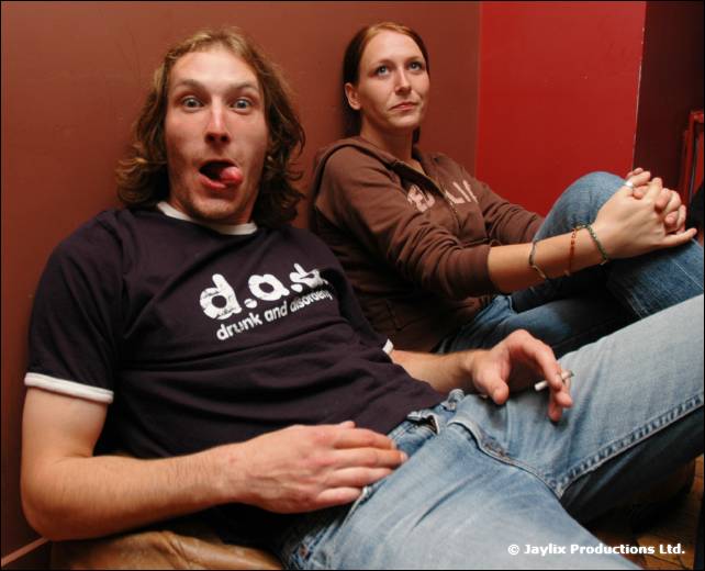 A man in a d.a.s. t-shirt sticks out his tongue while a woman sits behind him