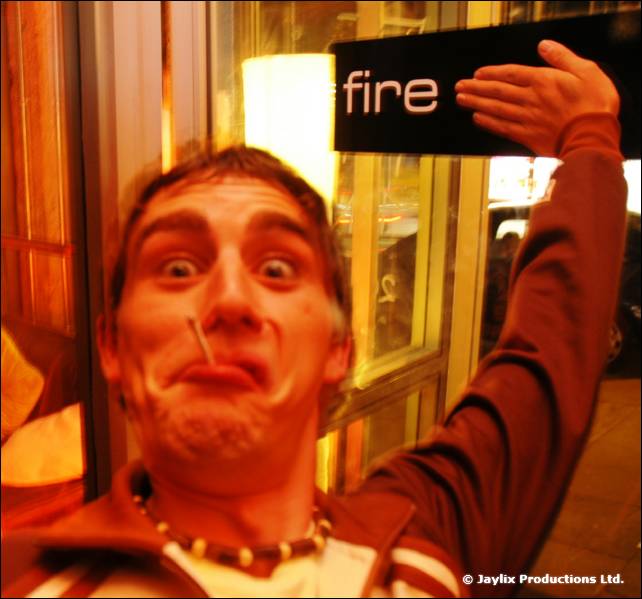A person with a cigarette in their mouth pulls a face and raises a hand beside a fire sign