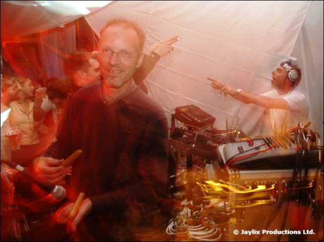 A smiling man in a dark jumper stands in a white tent with a DJ in headphones behind him
