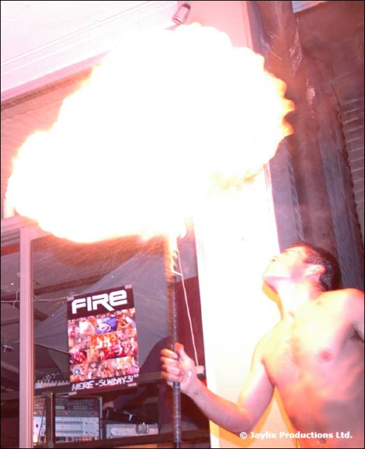 A shirtless person blows a huge fireball beside a FIRE poster