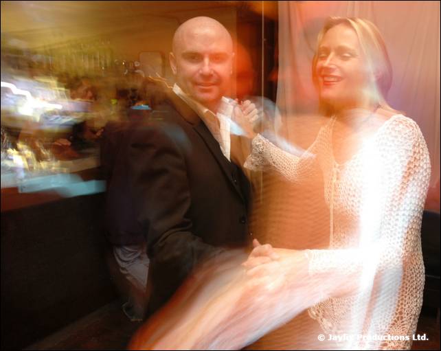 A bald man in a dark jacket and a woman in a white knitted top dance together, blurred