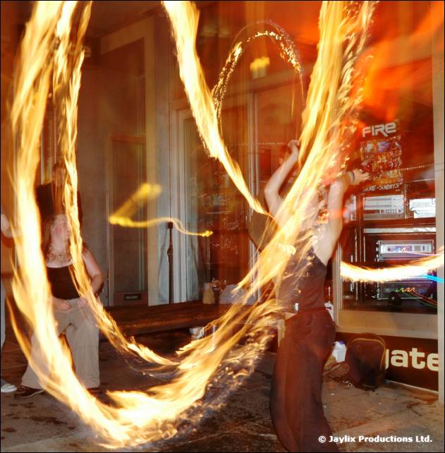 A person spins long trails of fire poi in a room with a gate sign and a FIRE poster behind