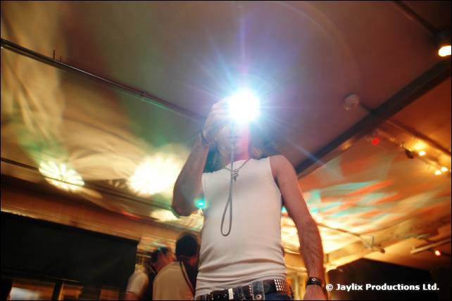 A person in a white vest holds up a camera with a bright flash, pointed at the lens