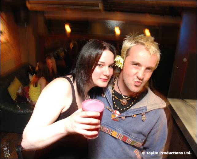 A woman holding a pink drink and a man with spiky blond hair pull faces at the camera