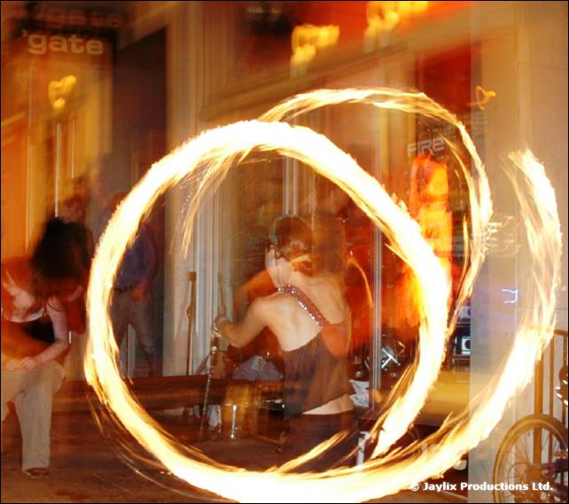 A large ring of fire poi trails in front of a bar window with a gate sign, a person behind