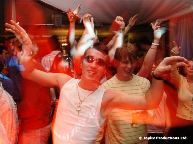 Crowd dancing with arms in the air, a man in sunglasses and a white vest at the front