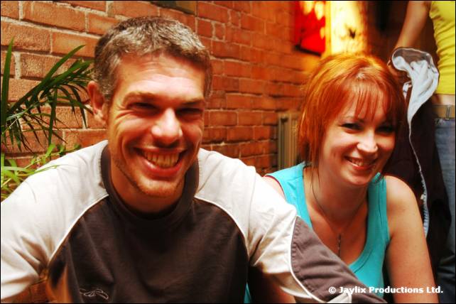 A smiling man and a woman with red hair sit close together in front of a brick wall