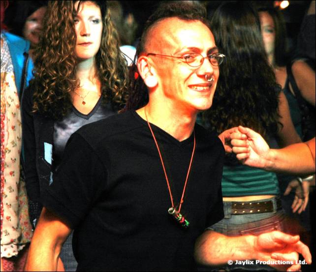 A smiling man with a mohawk and glasses wears a black t-shirt in a crowd