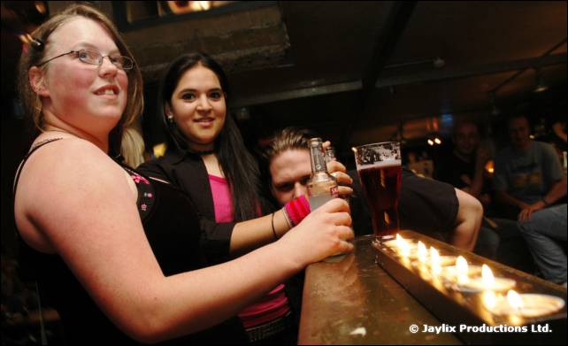 A woman in glasses holds a drink at a bar with a row of candles, two people behind