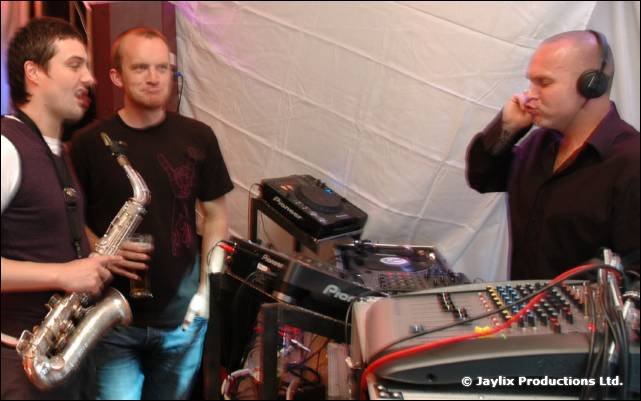 A saxophonist and another man stand next to a DJ in headphones at Pioneer decks in a white tent