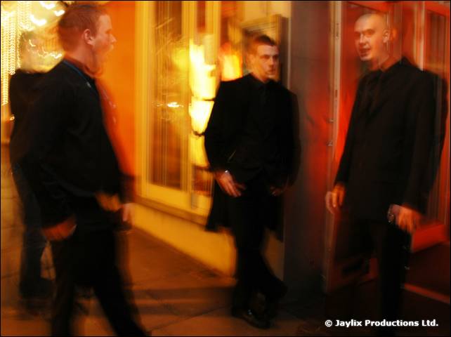 Three men in black clothes stand outside a lit doorway, blurred by movement