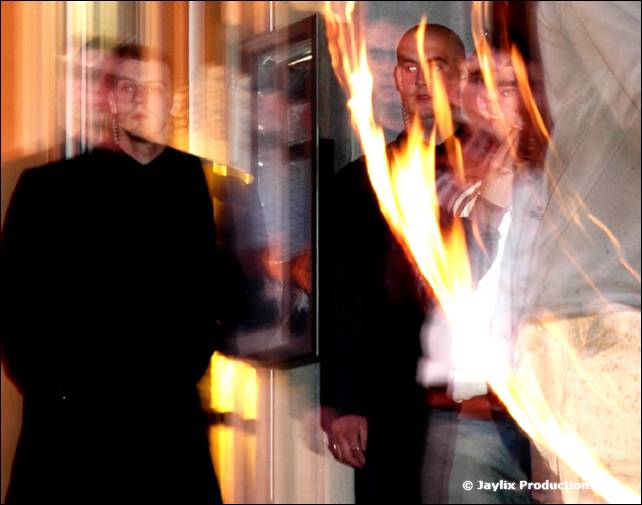 Two men in black stand at a window with a blurred flame at the right