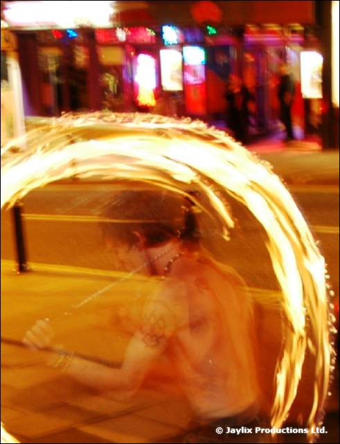 A blurred person behind a large circle of fire poi trails on a street at night