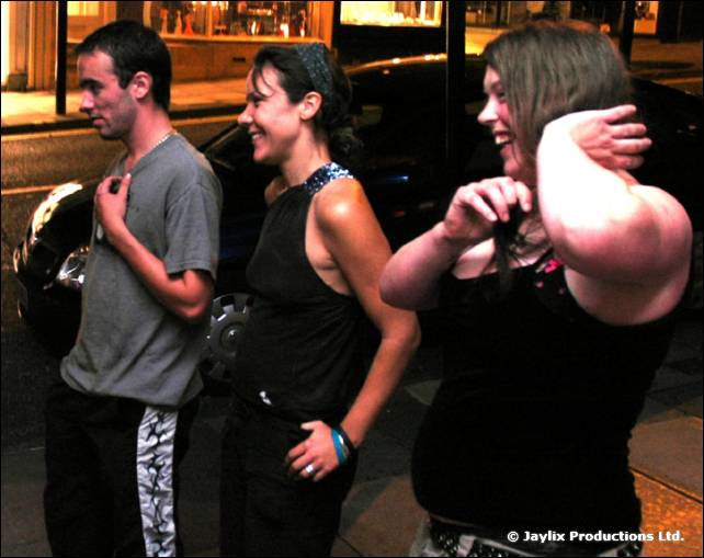 Three people stand laughing on a pavement at night, one covering their face with an arm