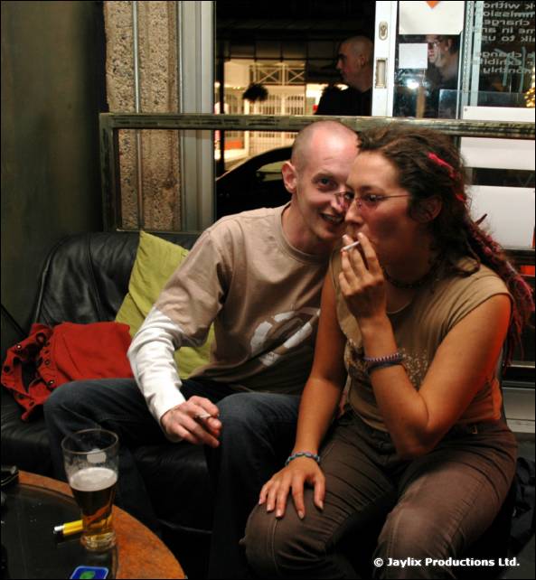 A bald man and a woman with dreadlocks smoke a cigarette on a leather sofa by a window