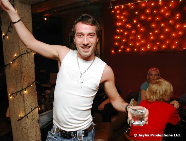A smiling man in a white vest holds a glass and leans on a pillar wrapped in fairy lights