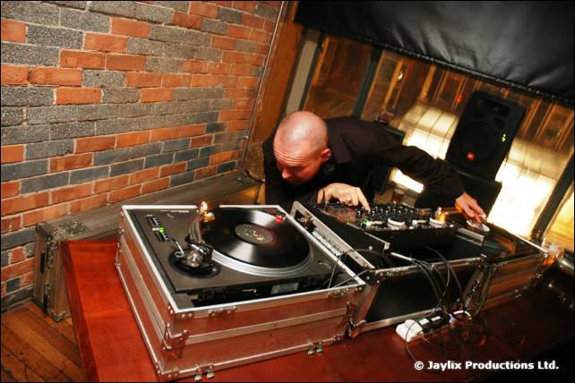 A bald DJ plays vinyl on a turntable in a flight case against a brick wall