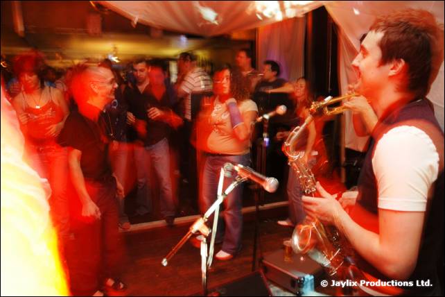 A saxophonist plays on the right in front of a blurred dancing crowd under white drapes