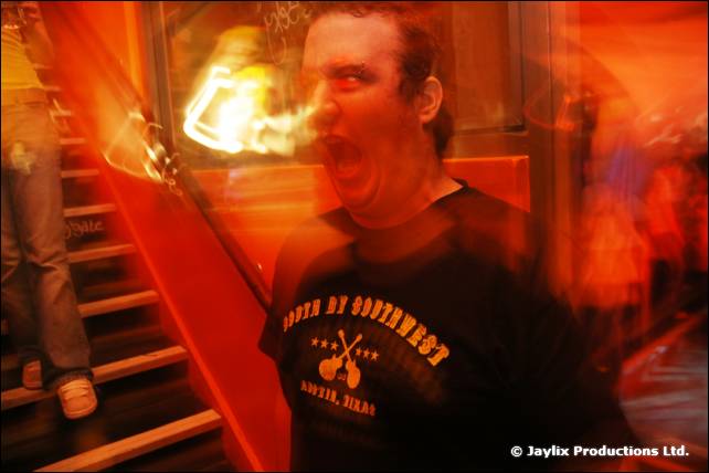A man in a black t-shirt has his mouth open with a flame blurred near his face, a staircase behind