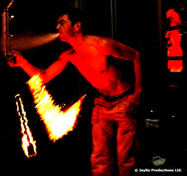 A shirtless person in red trousers blows fire from a torch in the dark