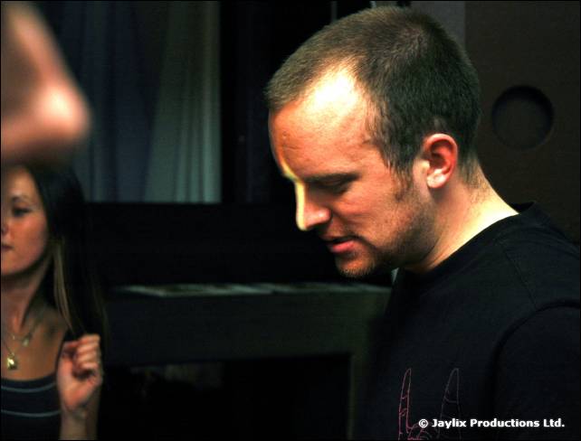 A man in a black t-shirt looks down, with a woman partly visible beside him