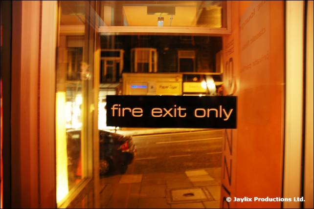 A lit sign reading fire exit only in a window looking onto a street at night