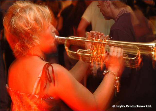 A blonde woman plays a trumpet in a red-lit bar