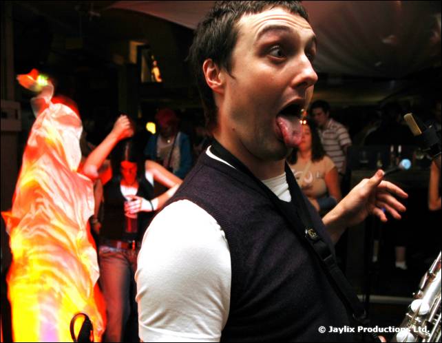 A man in a dark waistcoat and white top sticks out his tongue, a saxophone at the edge, dancers behind