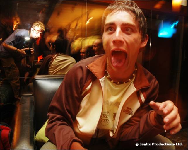 A man in a brown tracksuit top leans forward with his mouth open and points at the camera
