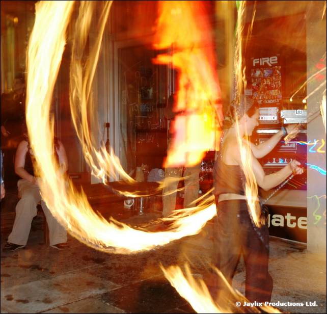 A person spins trails of fire poi in a room with a FIRE poster and a gate sign behind