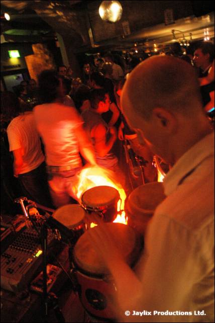 A man in a white shirt plays congas, with a flame glowing behind the drums and a crowd behind