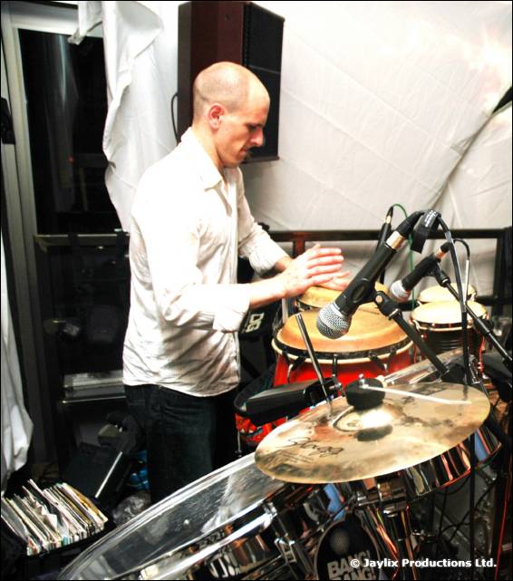 A bald man in a white shirt plays a percussion kit with microphones and cymbals inside a white tent
