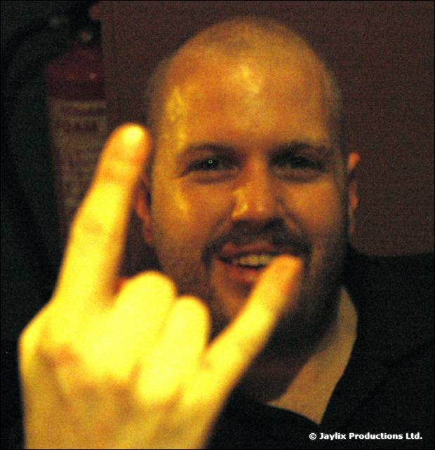 A smiling bald man with a beard makes a rock horns sign towards the camera
