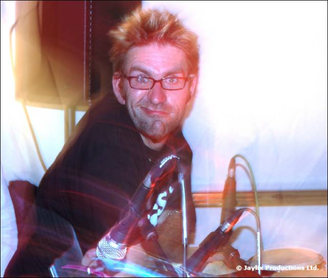 A smiling man with spiky hair and glasses sits by percussion and microphones