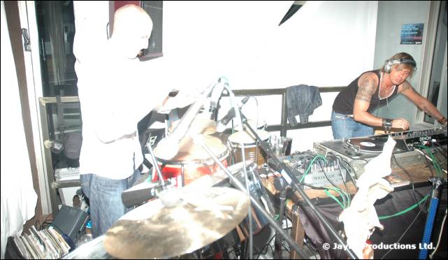 A man plays percussion on the left while a tattooed DJ in headphones mixes on the right, in a white room