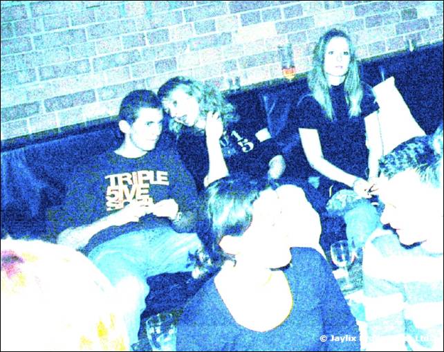 A group of people sit on a dark sofa by a brick wall, one wearing a Triple Five Soul t-shirt, in a blue-tinted shot