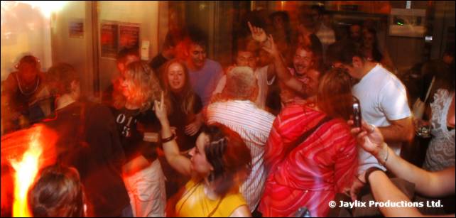 A crowd dances in a bar, with a person in a red striped hoodie in the foreground and a flame at the left